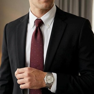 Puede incluir: Un traje negro con camisa blanca y corbata burdeos con lunares blancos. Se ve un reloj de pulsera plateado en la muñeca. La imagen es un primer plano de la parte superior del cuerpo, centrándose en el traje y los accesorios.