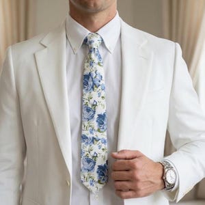 Peut inclure: Un costume blanc avec une chemise blanche et une cravate à motifs floraux. La cravate présente un motif de roses bleues sur fond blanc. Une montre-bracelet argentée est visible.