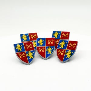 Puede incluir: Un conjunto de cinco pines con forma de escudo y diseño cuartelado. Cada pin muestra cuadrantes alternos en azul y rojo con figuras de leones amarillos y llaves cruzadas, todo ello delineado con un borde gris claro sobre fondo blanco.