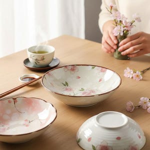 以下が含まれることがあります： ピンクと白の桜の花柄が特徴の、日本の陶器のボウルと湯呑みのコレクション。 ボウルは茶色の縁があり、箸と小皿とともに木製のテーブルに配置されています。