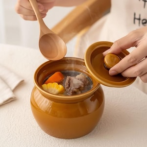 以下が含まれることがあります： 野菜と肉が入ったスープが入った、ふた付きの茶色のセラミック製の調理鍋。木のスプーンが鍋の上に置かれています。鍋は丸みを帯びた形状で、光沢のある仕上がりです。