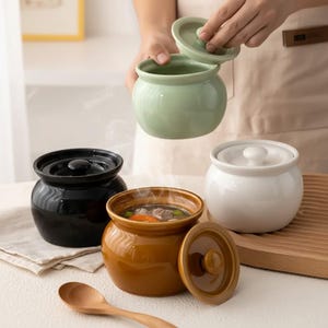 Puede incluir: Un conjunto de cuencos de cerámica para sopa en negro, blanco, marrón y verde. Un cuenco está abierto, revelando sopa humeante con verduras. Una cuchara de madera está cerca. Los cuencos tienen tapas y forma redondeada.