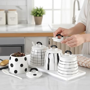 May include: A collection of white ceramic kitchen canisters with black accents. The set includes jars with polka dots, stripes, and a grid pattern, each with a black handle on the lid. A white tray holds the canisters, and a spoon rests nearby.