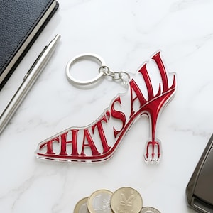 May include: A clear acrylic keychain in the shape of a high-heeled shoe with the words "THAT'S ALL" in red. The heel features a trident design. The keychain is on a silver ring, next to a black notebook, pen, coins, and a car key.