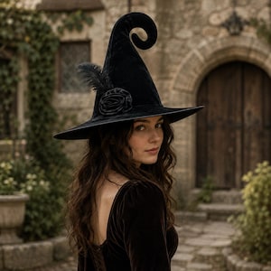 May include: A black velvet witch hat featuring a tall, curled pointed crown and a wide brim. The hat is adorned with a black fabric rose and a single black feather accent attached to the side, creating a textured and dark aesthetic piece.