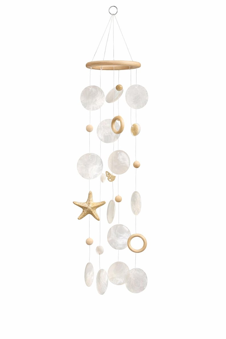 May include: A coastal-themed wind chime featuring a wooden ring and various shapes, including round, oval, and starfish. The chime includes white, pearlescent discs, wooden beads, and a small butterfly accent, all suspended by thin strings.