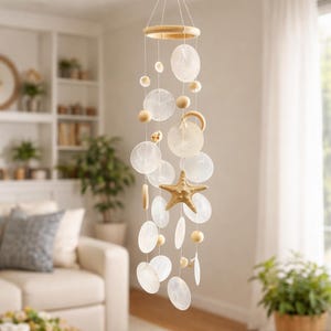 May include: A coastal-themed wind chime featuring a wooden ring and starfish accent. The chime has a series of white, shell-like discs and wooden beads suspended by strings. The starfish is a light brown color. The overall design is intended for indoor use.
