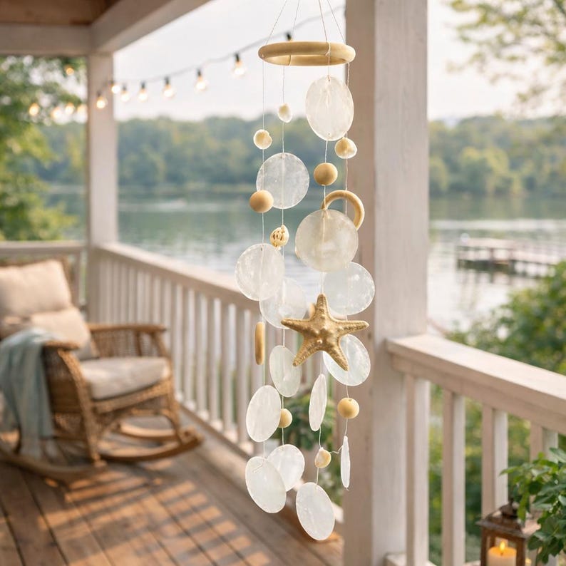 May include: A coastal-themed wind chime with a wooden ring and starfish accent. The chime features translucent, round shell discs and wooden beads, creating a gentle, decorative hanging ornament. The background shows a porch overlooking a lake.