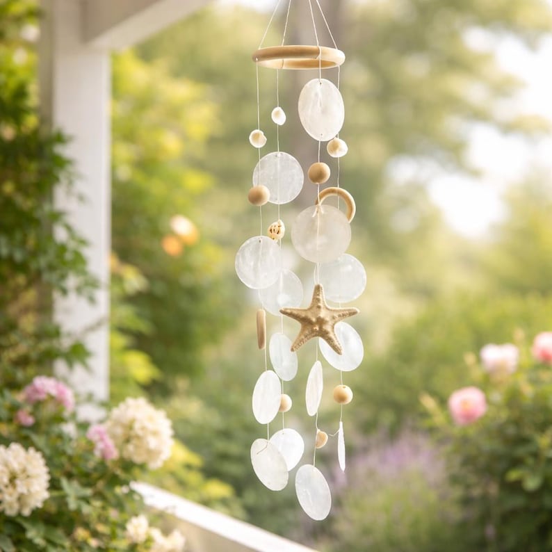 May include: A coastal-themed wind chime featuring a wooden ring and a starfish. The chime includes round, flat, and shell-shaped elements in white and beige, suspended by strings. Wooden beads and a starfish add to the design.