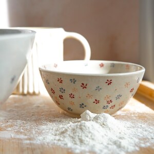 Puede incluir: Un cuenco de cerámica con un estampado floral en rojo, azul y naranja descansa sobre una superficie de madera espolvoreada con harina. Una taza blanca y otro cuenco se ven en el fondo, sugiriendo una escena de repostería.