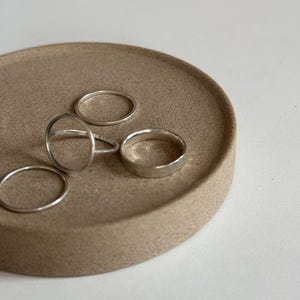 Puede incluir: Un conjunto de anillos de plata de varios diseños descansa sobre una bandeja circular beige. Los anillos incluyen una banda simple, un anillo con un diseño circular y dos bandas finas. La bandeja tiene una superficie texturizada.