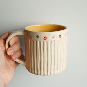 Puede incluir: Taza de cerámica color crema con textura acanalada vertical. El borde está decorado con pequeños patrones florales coloridos en rojo, azul y amarillo. El interior de la taza es de color amarillo cálido y tiene un asa a juego.