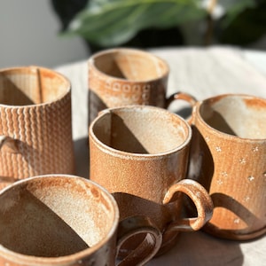 Op de afbeelding: Een verzameling handgemaakte keramische mokken in verschillende bruintinten. De mokken hebben een getextureerd oppervlak met patronen en stippen. Elke mok heeft een gebogen handvat.