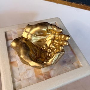 Könnte beinhalten: Eine goldfarbene Dekoschale in Form von zwei geformten Händen. Die Skulptur hat eine metallische Oberfläche mit detaillierten Finger- und Handflächenkonturen und bildet eine hohle Mitte, die zur Aufbewahrung kleiner Gegenstände dient.