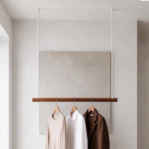May include: A ceiling-mounted clothing rack featuring a horizontal dark brown wooden rod suspended by two white ropes. Three wooden hangers are attached to the rod, holding a beige long-sleeve shirt, a white collared shirt, and a brown blazer.