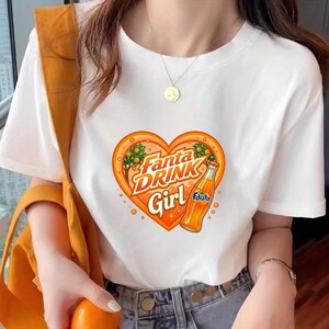 May include: A white short-sleeved t-shirt featuring a graphic of an orange heart containing the text Fanta DRINK Girl and an illustration of a Fanta orange soda bottle. The design includes small orange trees and sparkles within the heart shape.