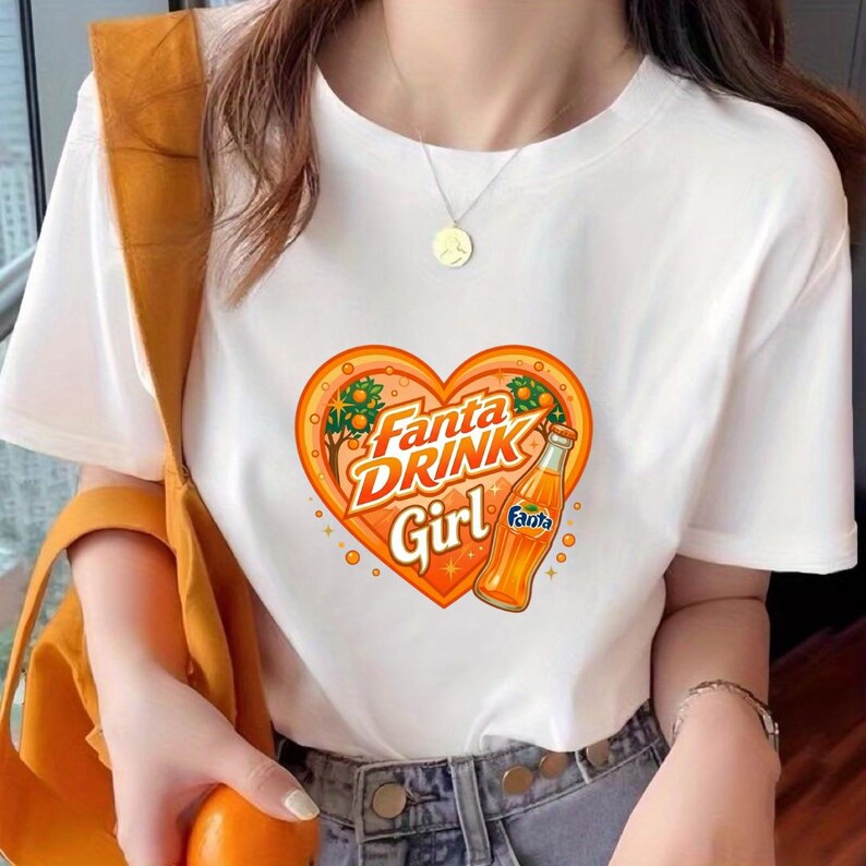 May include: A white short-sleeved t-shirt featuring a graphic of an orange heart containing the text Fanta DRINK Girl and an illustration of a Fanta orange soda bottle. The design includes small orange trees and sparkles within the heart shape.