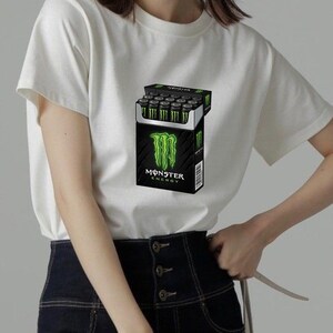 May include: A white short-sleeved t-shirt featuring a graphic print of a black rectangular box resembling a cigarette pack, filled with black cans with green claw logos. The box displays the text Monster Energy in white and green lettering.