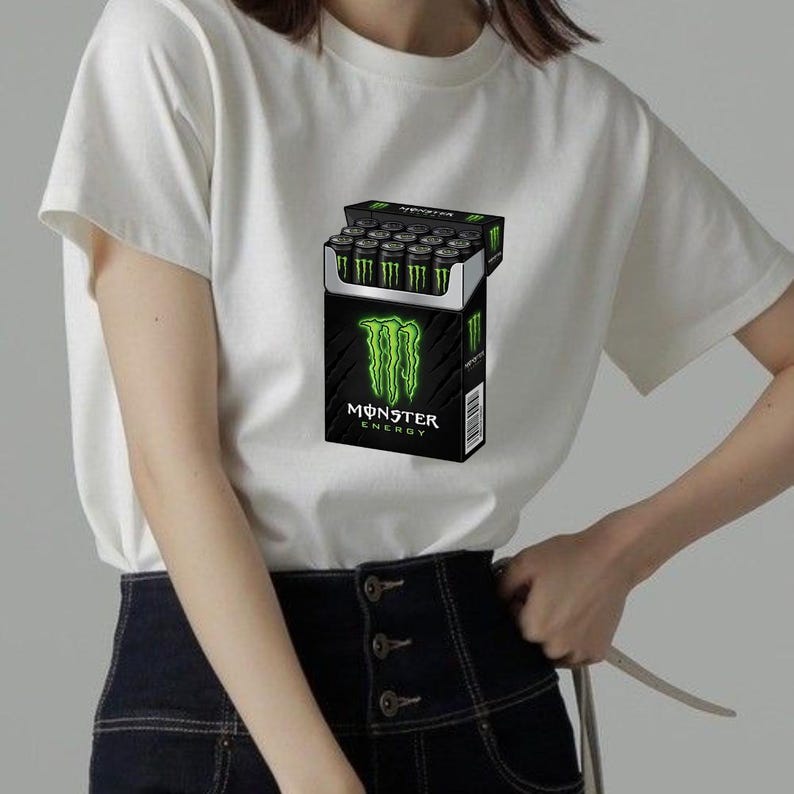 May include: A white short-sleeved t-shirt featuring a graphic print of a black rectangular box resembling a cigarette pack, filled with black cans with green claw logos. The box displays the text Monster Energy in white and green lettering.