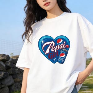 May include: A white short-sleeved t-shirt featuring a graphic print of a blue heart shape containing the text Pepsi Girl in white and red script. The design includes a small Pepsi soda can illustration with the Pepsi logo and sparkling star accents.