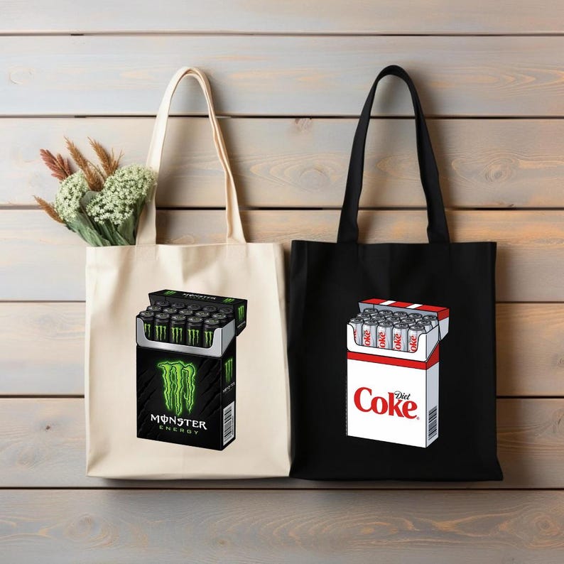 May include: A cream-colored canvas tote bag featuring a graphic of a Monster Energy drink pack and a black canvas tote bag featuring a graphic of a Diet Coke drink pack. The Monster bag has a small bouquet of dried greenery tucked inside the top.
