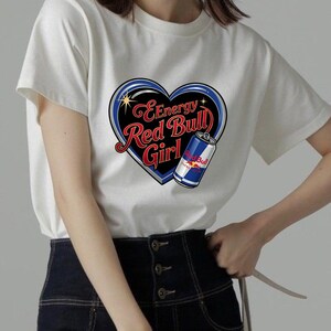 May include: A white short-sleeved t-shirt featuring a graphic of a black heart with a blue outline. Inside the heart, the text reads Energy Red Bull Girl in red and white script. A blue and silver Red Bull energy drink can is positioned on the right.
