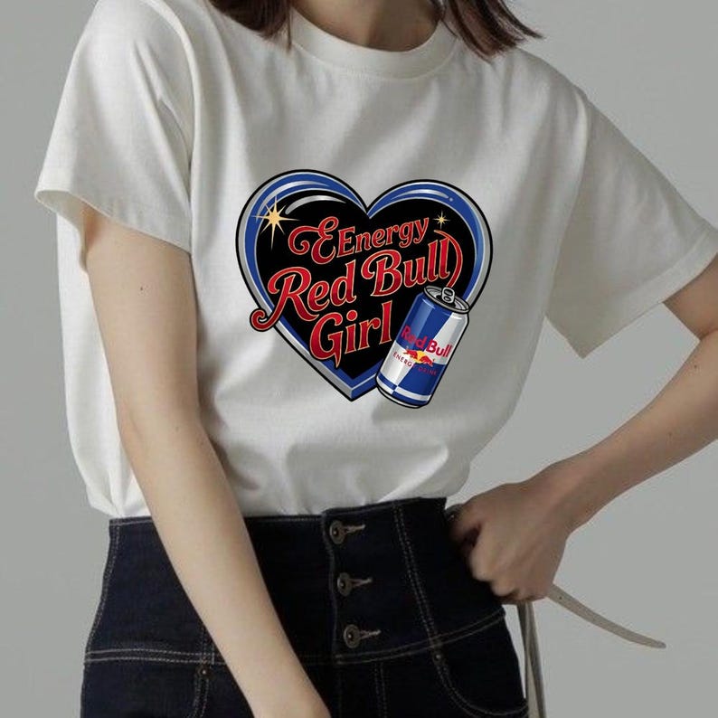 May include: A white short-sleeved t-shirt featuring a graphic of a black heart with a blue outline. Inside the heart, the text reads Energy Red Bull Girl in red and white script. A blue and silver Red Bull energy drink can is positioned on the right.