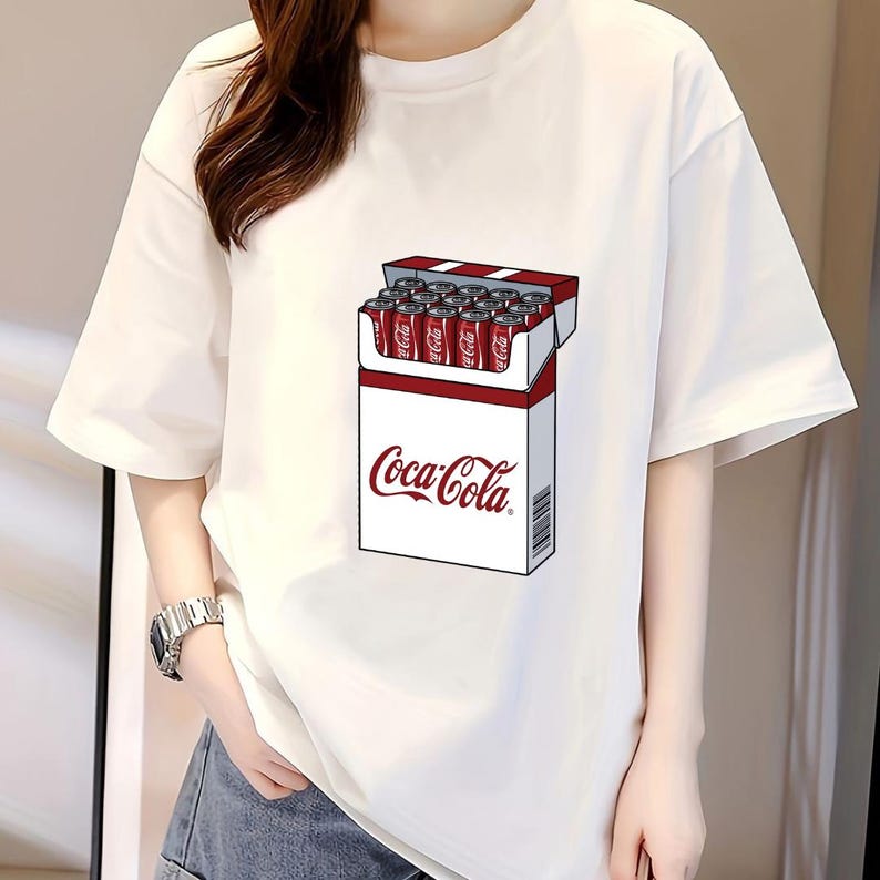 May include: A white short-sleeved t-shirt featuring a graphic print on the front. The graphic depicts a rectangular box resembling a cigarette pack, but filled with red Coca-Cola soda cans. The Coca-Cola logo is printed in red on the white box.