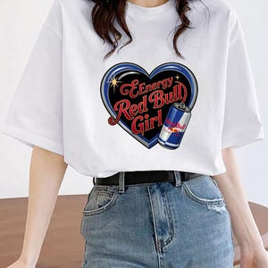 May include: A white short-sleeved t-shirt featuring a graphic of a black heart outlined in blue and silver. Inside the heart is the red text "Energy Red Bull Girl" and an illustration of a blue, silver, and red Red Bull energy drink can.