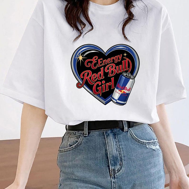May include: A white short-sleeved t-shirt featuring a graphic of a black heart outlined in blue and silver. Inside the heart is the red text "Energy Red Bull Girl" and an illustration of a blue, silver, and red Red Bull energy drink can.