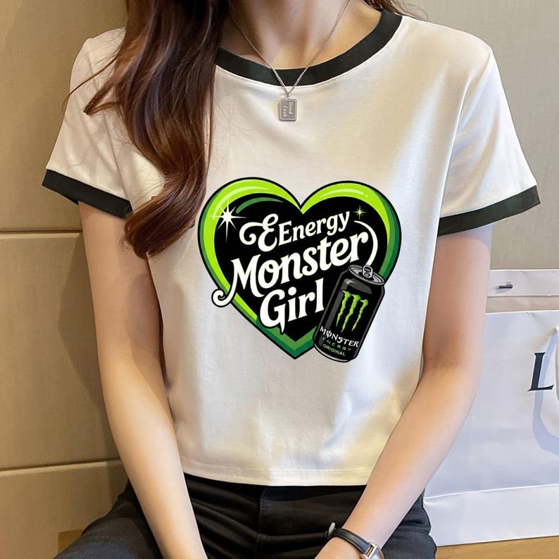 May include: A white short-sleeved t-shirt with black trim on the collar and sleeves. The front features a large green heart graphic containing the text Energy Monster Girl and an illustration of a black Monster Energy drink can.