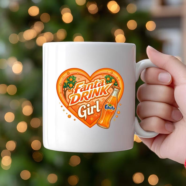May include: A white ceramic mug featuring a graphic of an orange heart containing the text Fanta DRINK Girl and an illustration of a Fanta orange soda bottle. The design includes small orange trees and sparkles within the heart shape.