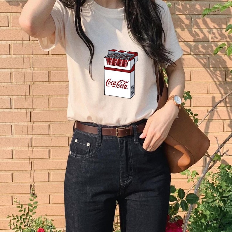 May include: A cream-colored short-sleeve t-shirt featuring a graphic print of a cigarette-style pack filled with red Coca-Cola cans. The pack is white with red accents and the Coca-Cola logo printed in red text on the front of the box.