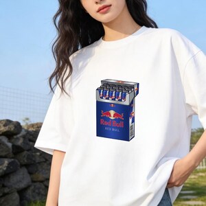 May include: A white short-sleeved t-shirt featuring a graphic print of a blue rectangular box designed to look like a cigarette pack, but filled with small blue and silver Red Bull energy drink cans, with the Red Bull logo and text printed on the front.