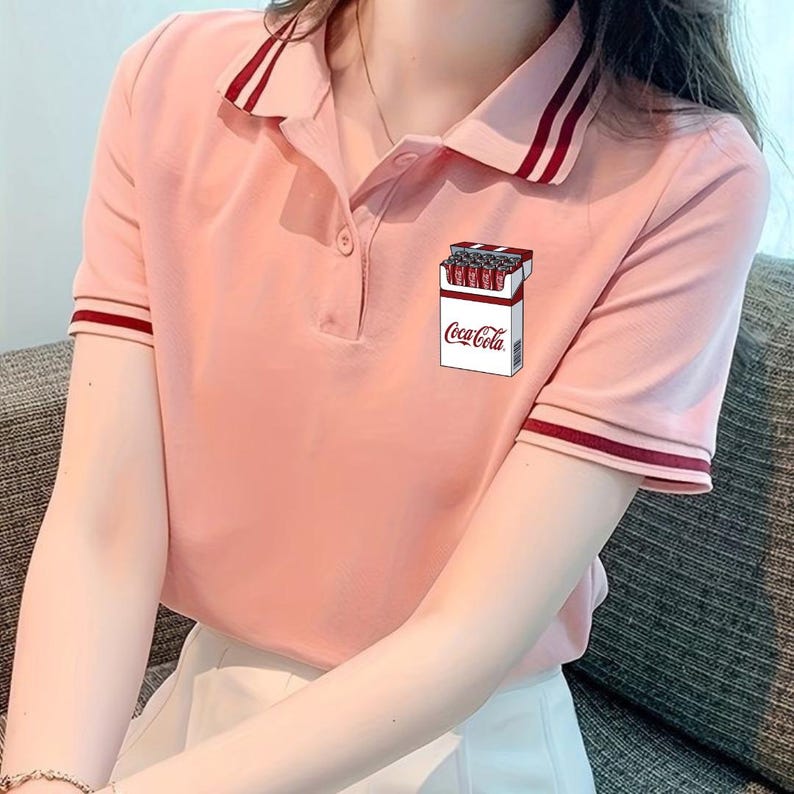 May include: A light pink short-sleeved polo shirt with a button-up collar featuring dark red striped trim on the collar and sleeve cuffs. A graphic print on the chest depicts a white rectangular pack containing red soda cans with the text Coca-Cola.