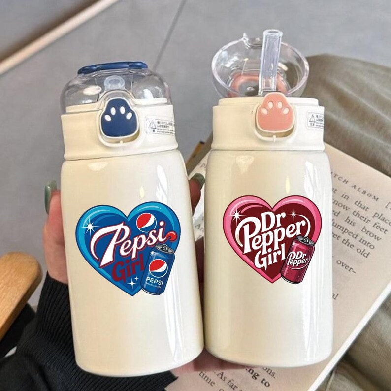 May include: Two off-white insulated water bottles with clear flip-top lids and paw-print buttons. The left bottle features a blue heart graphic with the text Pepsi Girl and a soda can. The right bottle has a pink heart with Dr Pepper Girl text.