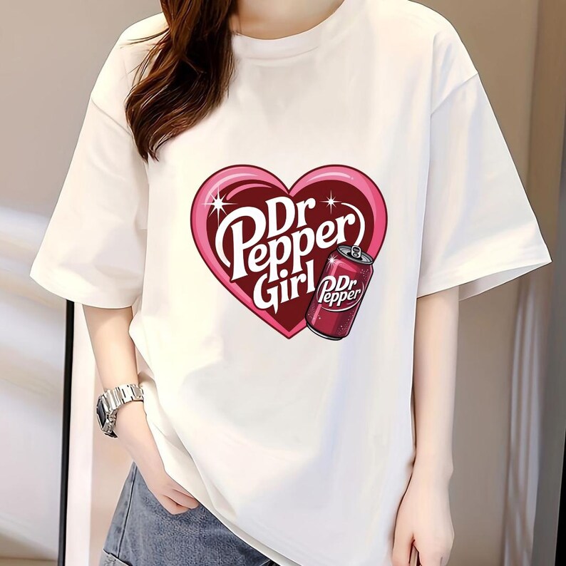 May include: A white short-sleeved t-shirt featuring a large pink heart graphic on the chest. Inside the heart, the text reads Dr Pepper Girl in white letters, accompanied by an illustration of a red Dr Pepper soda can with white text.