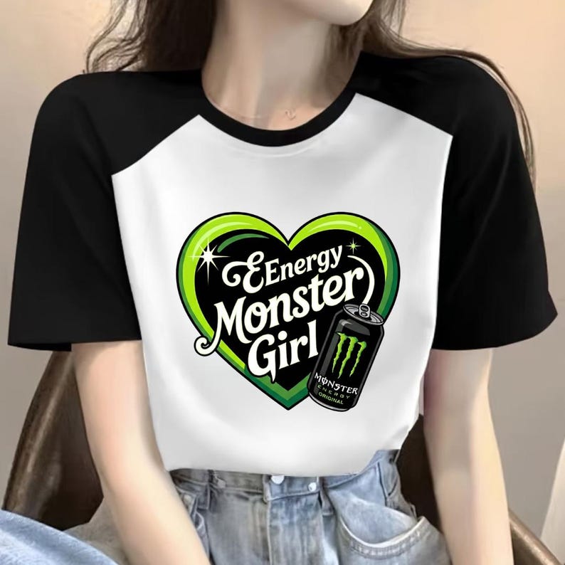 May include: A black and white raglan t-shirt featuring a graphic of a green heart with the text Energy Monster Girl and an illustration of a black Monster Energy drink can. The design is centered on the white front panel of the shirt.