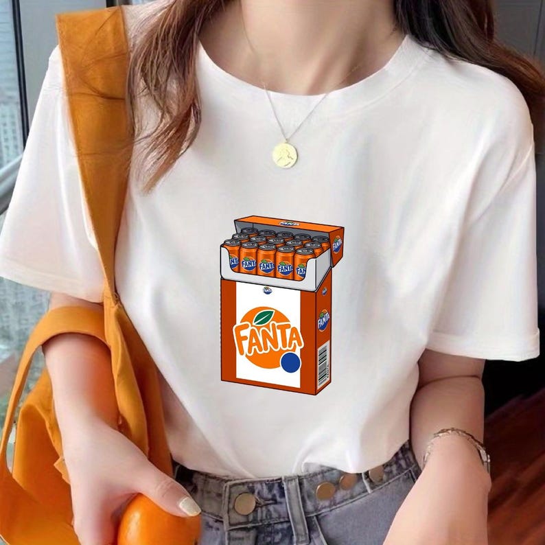 May include: A white short-sleeved t-shirt featuring a graphic print of an open orange box filled with Fanta soda cans. The box displays the Fanta logo in orange and white text with a green leaf icon, along with a barcode on the side panel.