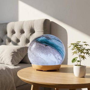 May include: A spherical lamp with a marbled blue, white, and grey design, set on a light wood base. The lamp is on a round wooden table next to a small potted plant. The lamp's design resembles a watercolour painting.
