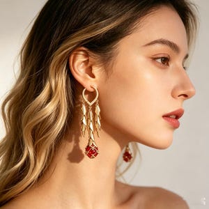 以下が含まれることがあります： ゴールドトーンのイヤリング、ティアドロップデザイン。 イヤリングは、ハート型のトップ、ぶら下がっている葉の形をしたアクセント、下部に赤い宝石のような石が特徴です。 長いウェーブのかかった髪の人が着用しています。