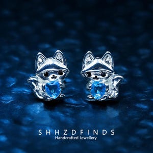 Puede incluir: Un par de pendientes de plata con forma de zorro, cada uno con una gema azul en forma de corazón. Los pendientes están sobre un fondo azul oscuro. El texto "SHHZD FINDS Handcrafted Jewellery" se muestra en la parte inferior.