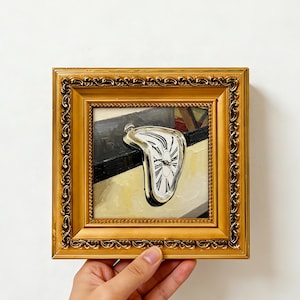 May include: A small square oil painting in an ornate gold frame depicting a melting pocket watch draped over a ledge. The watch face is white with black Roman numerals and hands. The background features a black and cream surface. Text reads SHHZD FINDS.