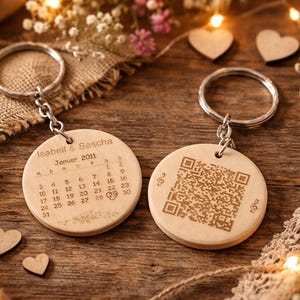 Könnte beinhalten: Zwei Holzschlüsselanhänger mit silbernen Ringen. Ein Schlüsselanhänger zeigt einen Kalender mit dem Text "Isabell & Sascha Januar 2011". Der andere Schlüsselanhänger hat einen QR-Code. Die Schlüsselanhänger liegen auf einer Holzoberfläche mit Dekorationselementen.