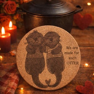 Könnte beinhalten: Ein runder Korkuntersetzer mit einem lasergravierten Design von zwei sich umarmenden Ottern, mit dem Text "We are made for each OTTER". Der Untersetzer befindet sich auf einer Holzoberfläche, mit Kerzen und einem gusseisernen Topf im Hintergrund.
