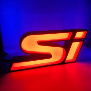 Peut inclure: Une enseigne lumineuse en trois dimensions représentant les lettres Si dans une police de caractères grasse et stylisée. Les lettres ont une bordure noire et émettent une lueur orange-rouge vive et chaude, créant un contraste marqué.