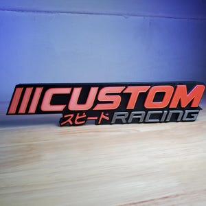 May include: A three-dimensional sign featuring the word CUSTOM in bold orange letters on a black background, with the Japanese text スピード and the word RACING in silver lettering positioned directly underneath the main text on the right side.