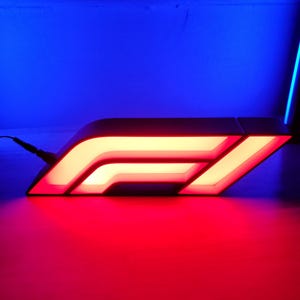 May include: A Formula 1 logo sign illuminated with bright red and white LED lights. The sign features the stylized F1 emblem with a black frame and a power cord extending from the left side, casting a vibrant red glow onto the surface below.