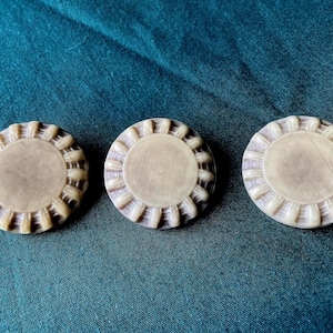 May include: Three round, off-white buttons with a decorative, ridged edge and a flat, circular center. The buttons are arranged on a dark teal fabric, creating a simple, yet elegant composition. The buttons appear to be made of a hard material.