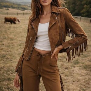 May include: A woman wearing a brown suede jacket with fringe detailing, a white tank top, and matching brown pants. The jacket has a zipper closure and the pants have a drawstring waist. The outfit is set against a backdrop of a field and rolling hills.
